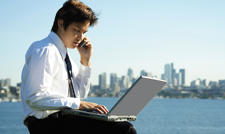 Man on a mobile phone with laptop on knees in front of a city skylines