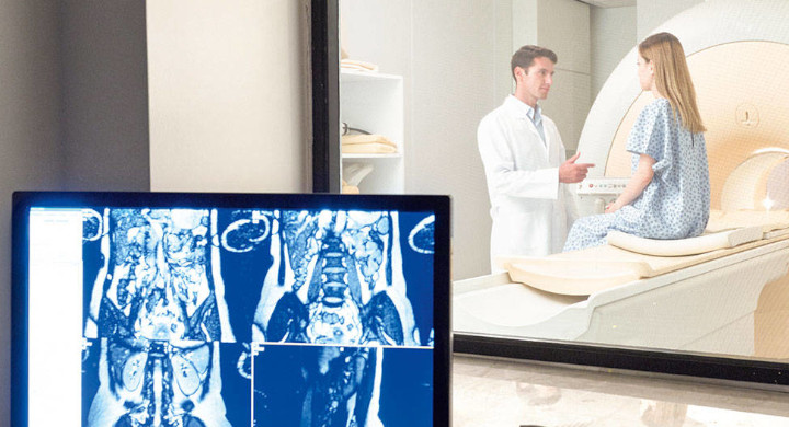 Male doctor talking to female patient sitting on MRI equipment in front of a display